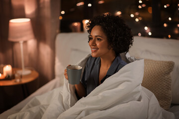 people, bedtime and rest concept - happy smiling woman in pajamas with coffee sitting in bed at...