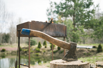 The ax is stuck in the stump. Grill and pond on background. Slow living in village.