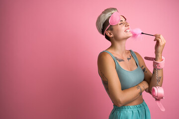 Overjoyed tattooed woman laughing with pink feather stick from sex shop on pink