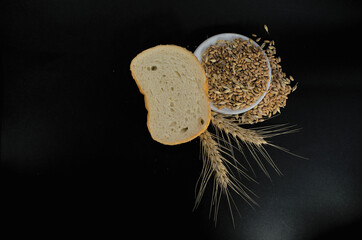 Grains of wheat on a plate next to white bread and ears of wheat on a black background