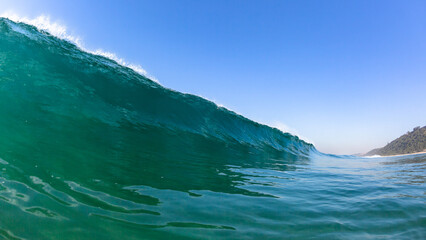 Ocean wave swimming close up encounter perspective of long sea wall of water about to crash break towards beach coastline with blue clear sky.