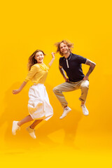 Portrait of young man and woman in casual clothes posing, cheerfully jumping isolated over yellow studio background