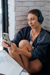 Young woman listening to music or podcast or lectures in wireless blue headphones while sitting near brick wall