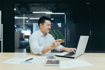 Plakat Cheerful and smiling asian businessman working in modern office, dark colors, making online purchase, using bank credit card and laptop, for online shopping