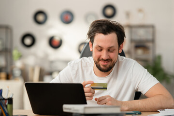 Cheerful young guy orders gift to his girlfriend. He prefers to shop online because it saves time. In front of him is laptop. He holds bank card and places an order.