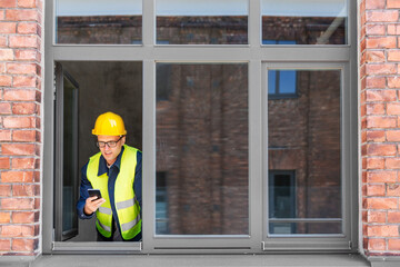 construction business and building concept - smiling male builder in helmet and safety west with smartphone at open window