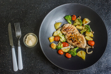 Grilled chicken breast, beef, steak, potatoes, cherry tomatoes, broccoli on a black plate, sauce cup and fork on black background.