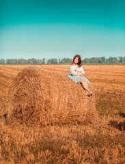 The girl in sits on a roll, stack of hay. The girl is walking in the field, it is a hot summer. The girl is wearing a blue skirt.