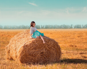 Naklejka na ściany i meble The girl in sits on a roll, stack of hay. The girl is walking in the field, it is a hot summer. The girl is wearing a blue skirt.