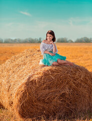 Naklejka na ściany i meble The girl in sits on a roll, stack of hay. The girl is walking in the field, it is a hot summer. The girl is wearing a blue skirt.
