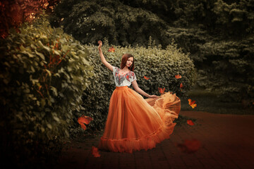 A girl in an orange skirt stands in the wind in an autumn park. Autumn yellow leaves fly around....
