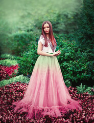 Fototapeta premium A slender girl stands among pink flowers in a long puffy dress. Around the lush foliage, many flowers, fabulous forest. The girl has long hair and makeup.