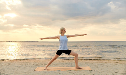 Fototapeta na wymiar fitness, sport, and healthy lifestyle concept - happy woman doing yoga warrior pose on beach over sunset