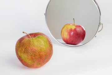 Old apple with a shriveled peel in front of a cosmetic mirror. Reflection of a ripe fresh apple in the mirror. Metaphor of cosmetology anti-aging procedures, skin care.