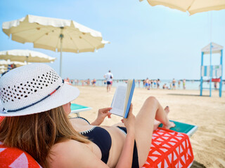 Woman wearing a summer hat and lying on a deckchair at the beach while relaxing reading a book, in the background umbrellas, a lifeguard tower and bathers cooling off by the Adriatic sea, Italy. - obrazy, fototapety, plakaty