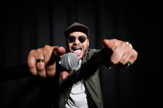 excited middle east hip hop performer in sunglasses and cap singing in microphone and gesturing on black.