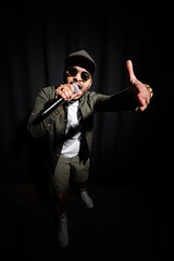 Fototapeta na wymiar emotional middle east hip hop performer in sunglasses and cap singing in microphone and gesturing on black.