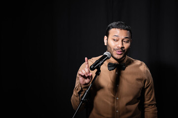 indian stand up comedian pointing with finger while telling jokes into microphone on stand on black background.