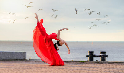 Dancing ballerina in a red flying skirt and leotard on the ocean embankment or on the sea beach surrounded by seagulls in the sky.