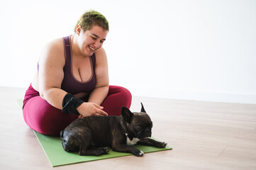 Happy young overweight woman exercising with a dog. Plus size female smiling while petting her French bulldog, sitting together on yoga mat in studio.