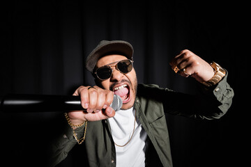 indian hip hop performer in sunglasses and cap singing loud in microphone on black. - obrazy, fototapety, plakaty