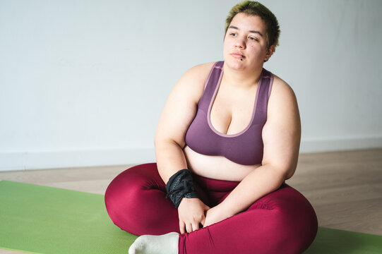 Tired overweight woman with melancholia sitting on yoga mat, getting motivation for a workout session. Obese Caucasian female in her 20s looking unhappy and pensive.