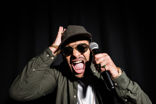 emotional middle east hip hop performer in sunglasses and cap screaming while holding microphone isolated on black.
