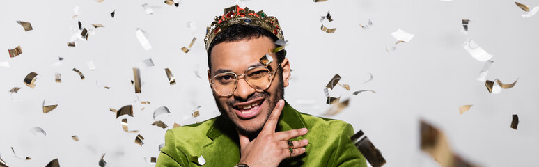 cheerful indian hip hop performer in green velvet blazer and crown near falling confetti on grey,...