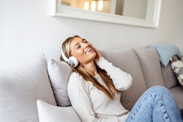 Happy millennial female with eyes closed in wireless headphones and casual clothes sitting alone on soft couch with hands behind head and listening with pleasure to favorite music 