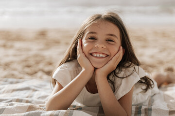 Delighted stylish kid with hands on cheeks relaxing on sandy seashore
