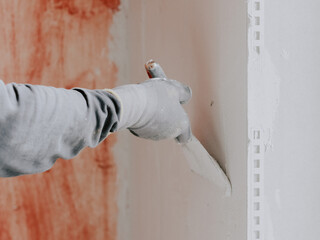 The hands of a young caucasian male plasterer in construction gloves apply fresh putty on the wall