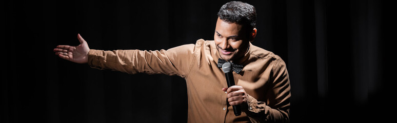 smiling indian comedian performing stand up comedy into microphone on black, banner.