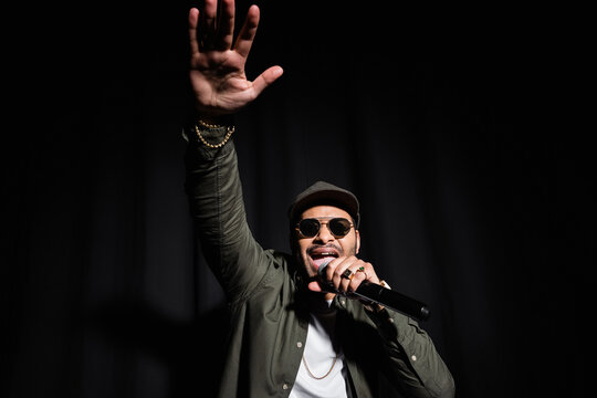 emotional middle east hip hop performer with raised hand singing in microphone on black.