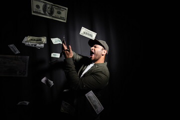 excited eastern hip hop performer in cap throwing dollar banknotes on black.