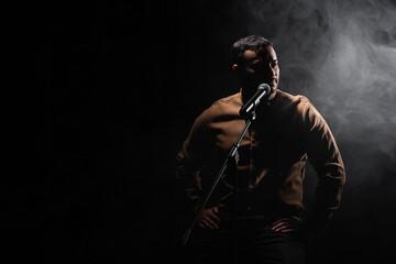indian comedian sitting on chair and performing stand up comedy into microphone on black with smoke.