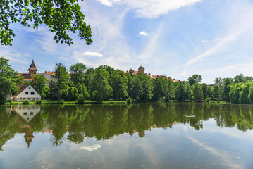 in the photo there is a pond surrounded by trees and buildings are displayed. The reflection of the world around is reflected from the surface. The sky is blue and it's a beautiful sunny day.