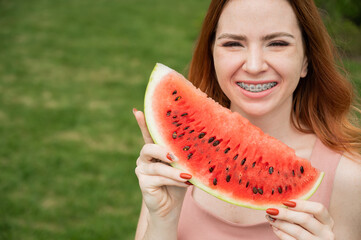 Beautiful red-haired woman smiling with braces and about to eat a slice of watermelon outdoors in summer