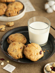 Oatmeal cookies with cranberry, cashew nuts and white chocolate near glass of milk on a metal tray. Brown table, plate of cookies on the background