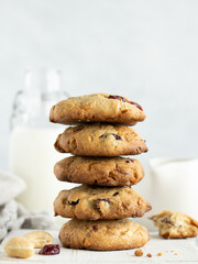 Close up stack of oatmeal cookies with cranberry, cashew nuts and white chocolate on a white wooden cutting board. Glass bottle of milk, kitchen towel and milk jug on the background