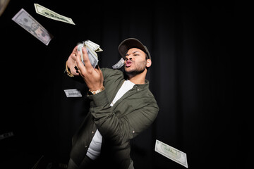 rich indian hip hop performer in cap throwing blurred dollar banknotes on black.