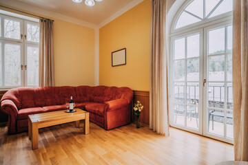Old hotel living room with red couch and wooden coffee table. In room can be found balcony window in arc shape, wooden floor and yellow-painted walls. There are also little additions as wine set.