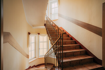 Hotel staircase with slightly curved architecture, windows and historic metal railing. Stairs are tiled and the handrail is supplemented with wooden handrail. There is also wood on walls and paneling.