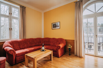 Old hotel living room with red couch and wooden coffee table. In room can be found balcony window in arc shape, wooden floor and yellow-painted walls. There are also little additions as wine set.