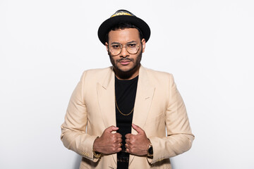 indian hip hop singer in fedora hat and eyeglasses adjusting blazer isolated on grey.