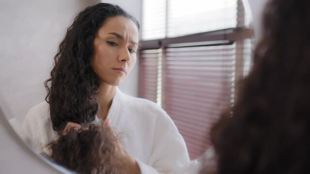 Worried upset young woman standing in bathroom in bathrobe reflected in mirror looking at split ends brittle damaged dry hair unhappy girl feeling frustration about unhealthy long curly fragile hair