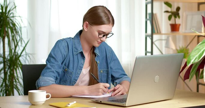A girl student in glasses uses a laptop and writes in a notebook while learning a lesson online in the interior. A young woman is being educated remotely in a room.