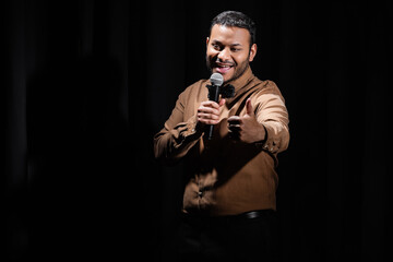 joyful middle east comedian showing like and performing stand up comedy into microphone isolated on black.