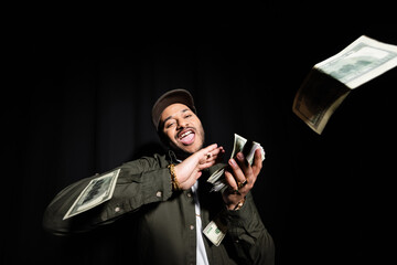 excited and rich indian hip hop performer in cap throwing dollar banknotes on black.