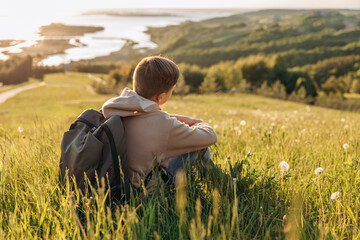 Tourist with backpack sitting on top of hill in grass field and enjoying beautiful landscape view. Rear view of teenage boy hiker resting in nature. Active lifestyle. Concept of local travel