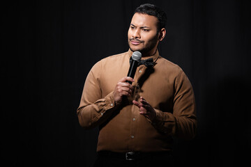 skeptical indian comedian in shirt and bow tie holding microphone during monologue on black.